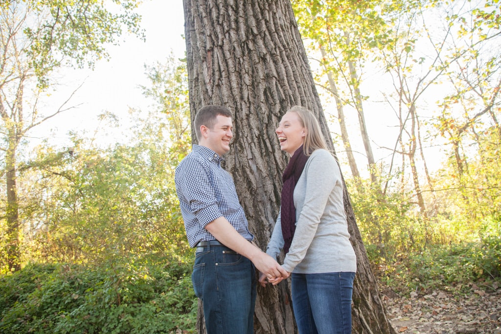 A newly engaged couple hold hands and laugh carelessly in front of a giant tree in the forest.