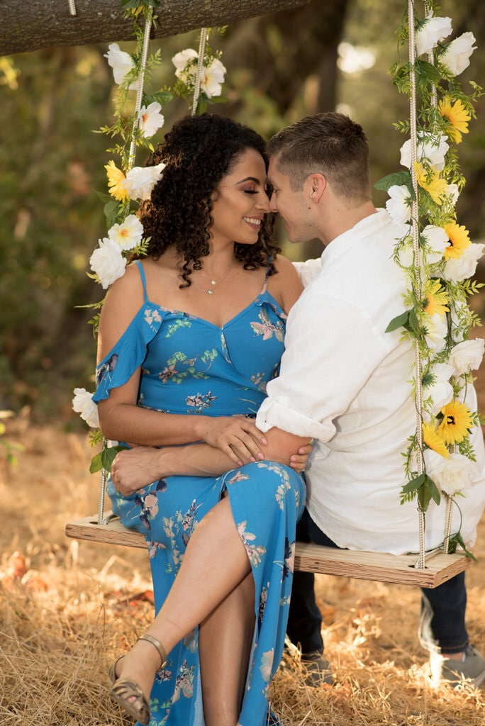 A recently engaged couple sit closely on a swing lined with fresh cut flowers. Their faces are close and they smile broadly and each other.