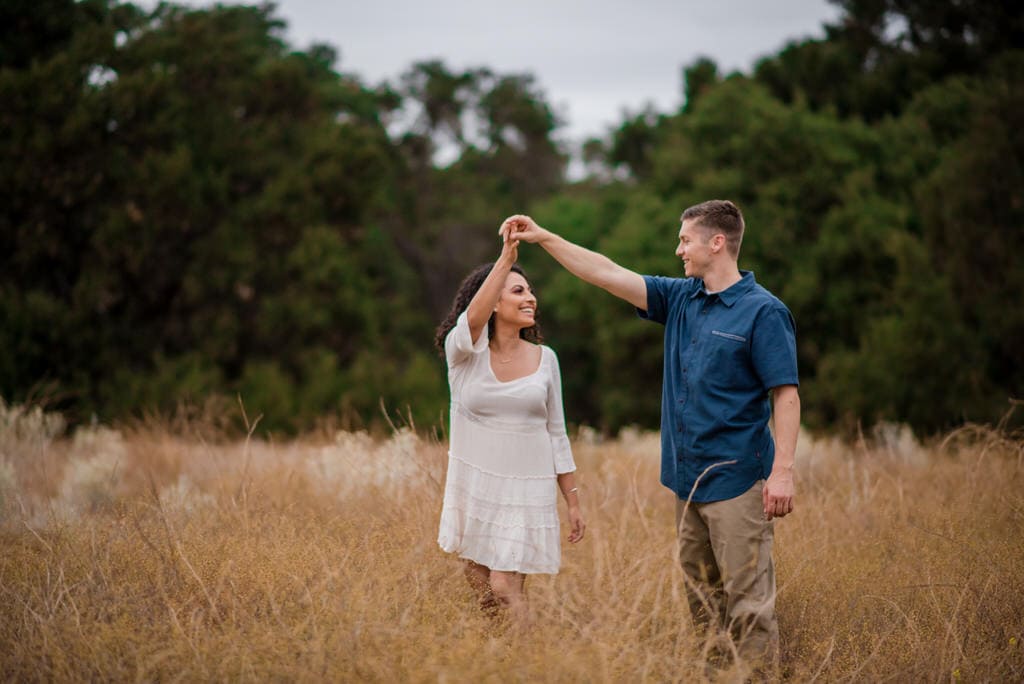 A man twirls his soon-to-be bride in the middle of a tall field surrounding by trees.