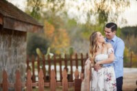 Cheyenne & Ken 6/9/18 A man grabs his soon-to-be wife around the waist as she smiles back at him. They leisurely lean against the fence of a cabin as they embrace each other.