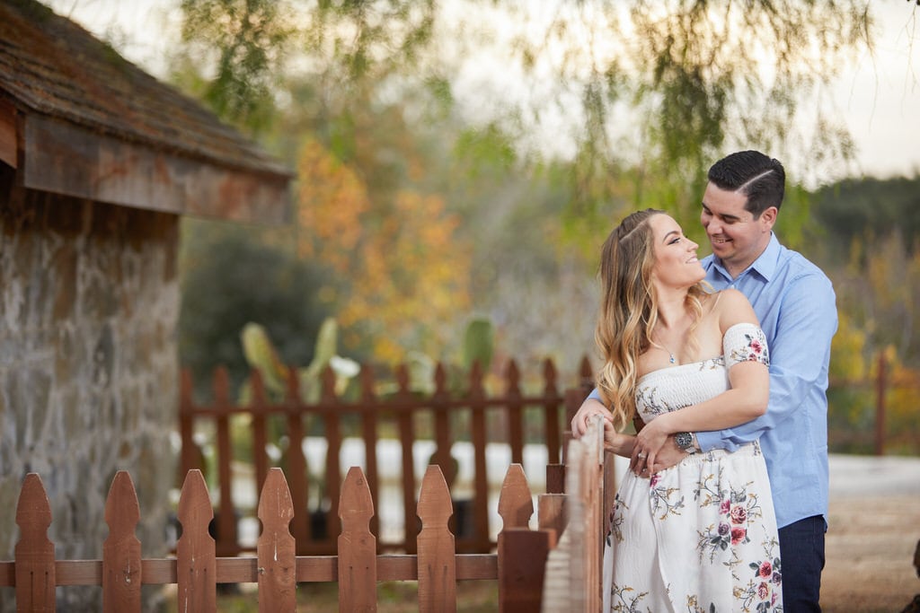 A man grabs his soon-to-be wife around the waist as she smiles back at him. They leisurely lean against the fence of a cabin as they embrace each other.