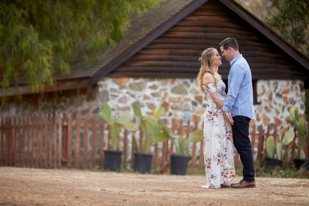 A couple touches hands and looks into each other's eyes in front of a stone cabin lined with cactuses.