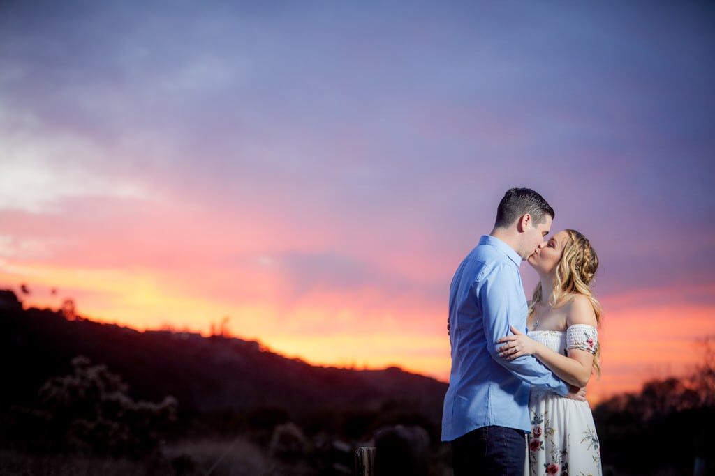 A young, newly engaged couple kiss sweetly as the sun sets in the background.