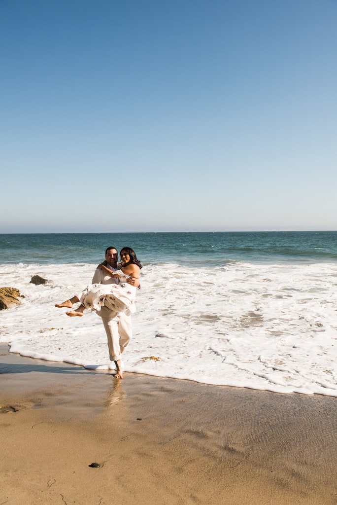 A man playfully carries his soon-to-be wife from the waters of the ocean.