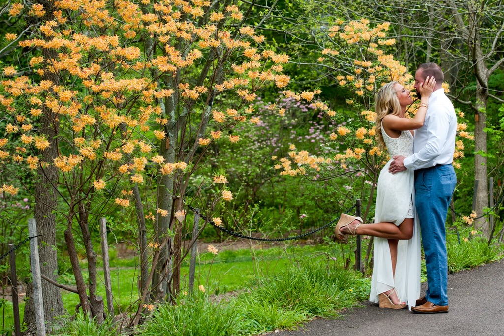 A couple stops on a path lined with blossoming trees to embrace each other. The woman's foot pops with excitement.