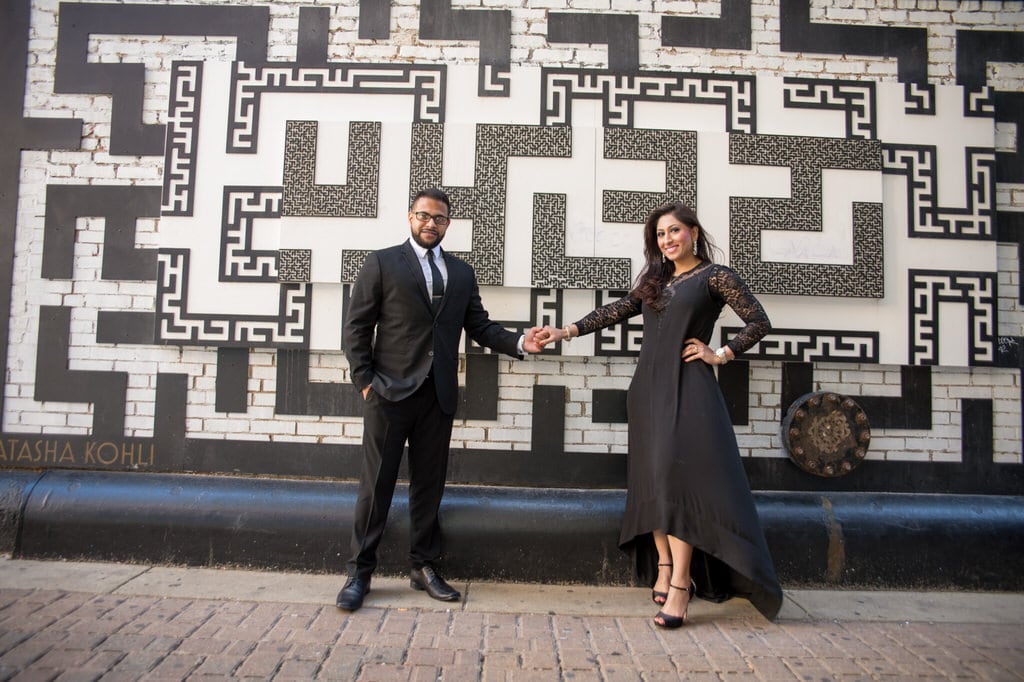 A newly engaged couple holding hands in front of a patterned wall outdoors.