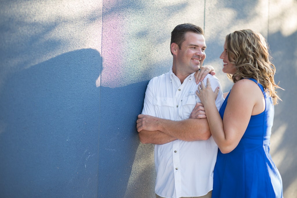 A couple looks at each other lovingly outdoors on a sunny day against a wall.