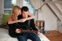 Wedding / Engagement Photography A woman admires her new fiance's guitar playing as they lounge on the end of their bed. She has her arms wrapped around his shoulders and they both smile thoughtfully as he plays.
