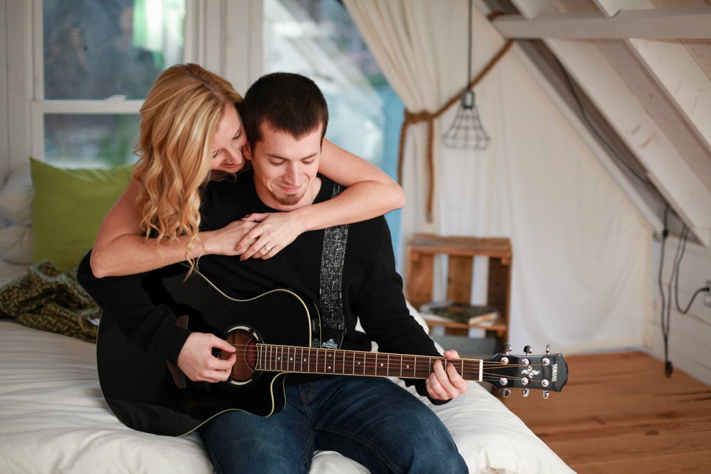 A woman admires her new fiance's guitar playing as they lounge on the end of their bed. She has her arms wrapped around his shoulders and they both smile thoughtfully as he plays.