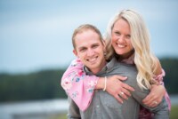 Hannah & Kevin 3/17/18 A newly engaged couple smile broadly at the photographer. The woman is on the man's back wearing a baby pink, floral bell sleeved dress.
