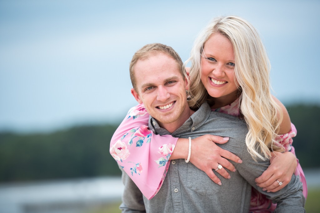 A newly engaged couple smile broadly at the photographer. The woman is on the man's back wearing a baby pink, floral bell sleeved dress.
