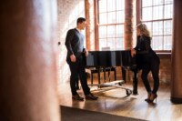 Wedding / Engagement Photography A stylish young couple shares a glance across a grand piano in an exposed brick loft with hardwood floors.