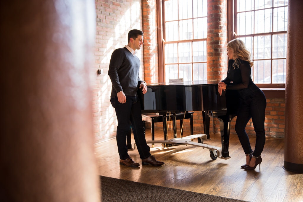 A stylish young couple shares a glance across a grand piano in an exposed brick loft with hardwood floors.