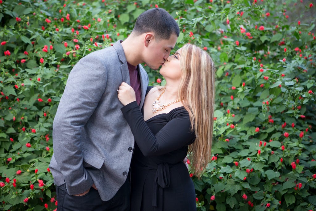 The young couple shares a firm kiss in front of high primrose bushes. The woman wears a classic black wrap dress with a statement necklace.