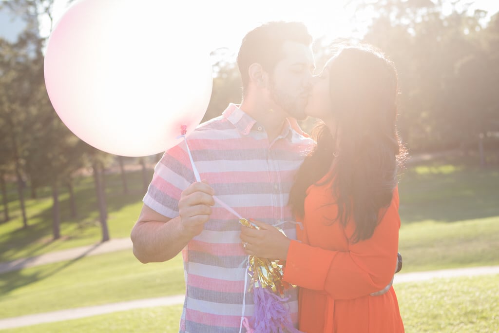 A young couple celebrates their engagement with balloons and streamers on a sunny day in the park.