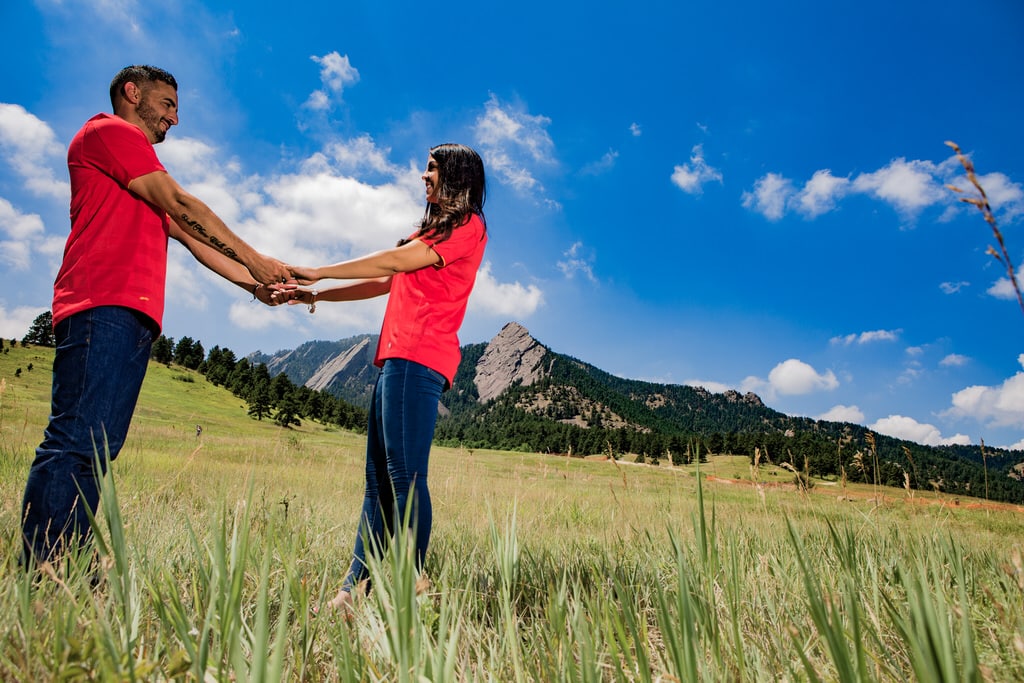 A young couple in matching red t-shirts and denim hold hands and gaze into each other's eyes. The beautiful landscape setting is adorned by gorgeous tree-covered mountains in the background.