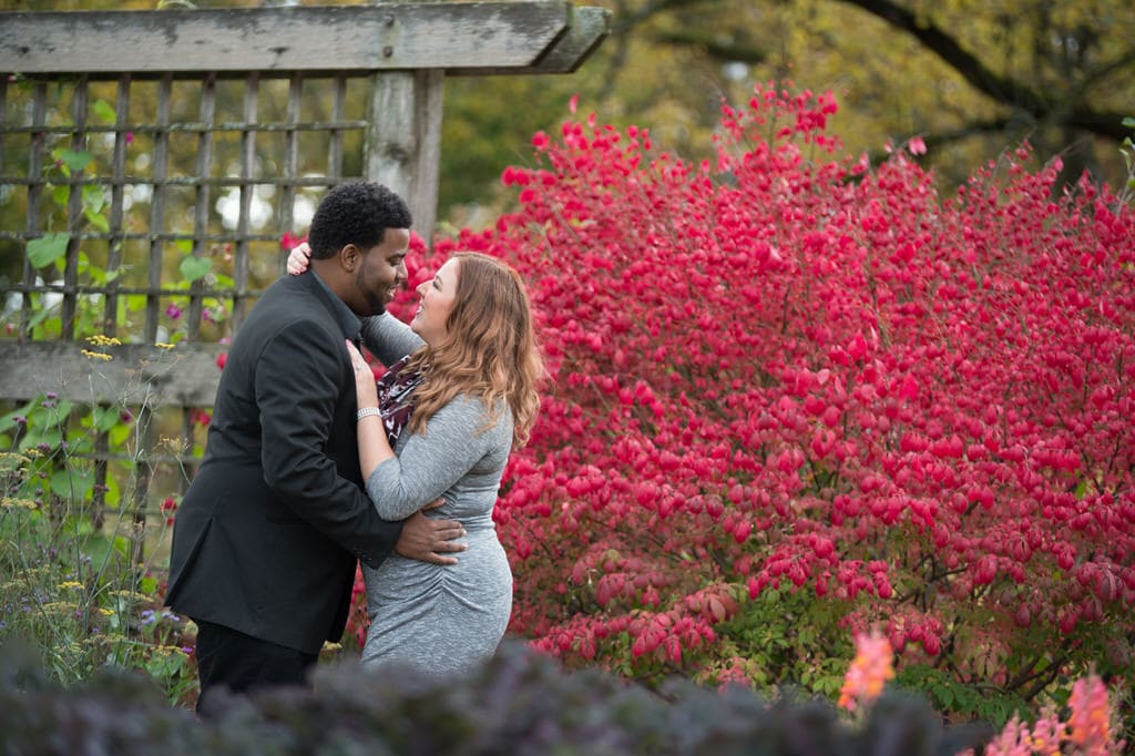 This couple holds one another while celebrating their fabulous engagement and time together while standing in front of a rustic wood garden wall and a beautiful vibrant bush.