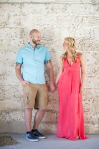 Melissa & Pat 9/9/17 A couple smiles at one another while holding hands against a stone brick wall.