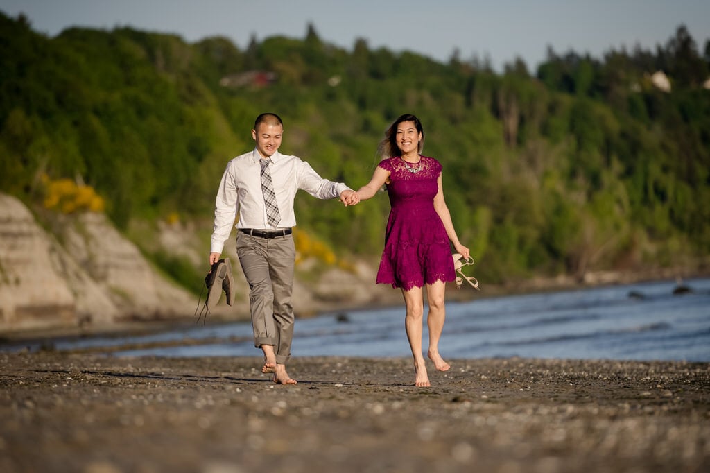 A light-hearted couple walks along the beach hand-in-hand as they smile at the photographer.