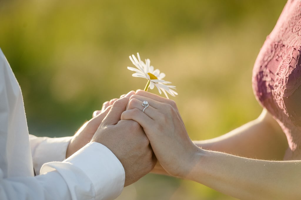 A couple holds a daisy between their hands and accent the diamond ring on her finger.