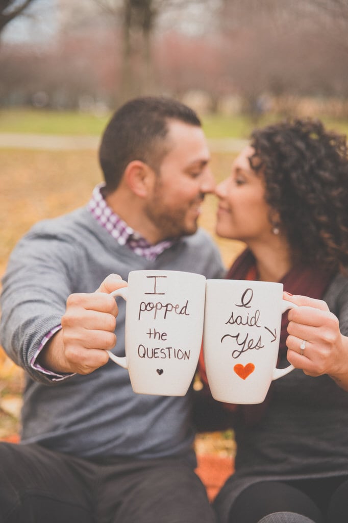 A young couple sits in the park and displays their matching coffee mugs advertising their new engagement.