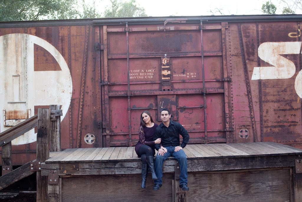 A newly engaged couple sits on a deck next to a freight train.