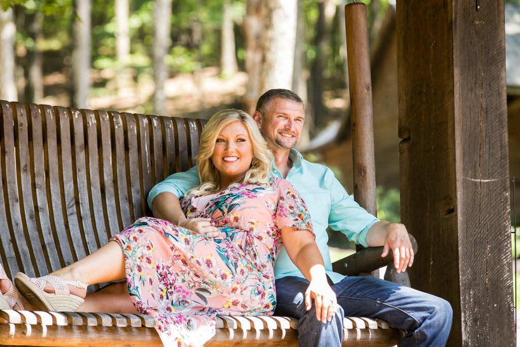 A newly engaged couple sits on a bench with broad smiles and bright eyes on a sunny day.