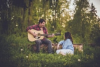 Shawnte & Justin Engagement A young couple locks eyes as the man plays his soon-to-be wife a song from his guitar. They are sitting on a log in the middle of the forest with dandelions scattered before them.