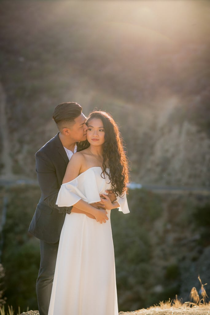 A newly engaged young couple takes portrait style photos in the mountain scenery of California. The man holds his fiance around the waist and kisses her cheek.