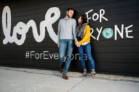 Sara & Joseph 9/15/18 A young couple holds hands and smiles adoringly at one another against a graphic painted wall on a city street.