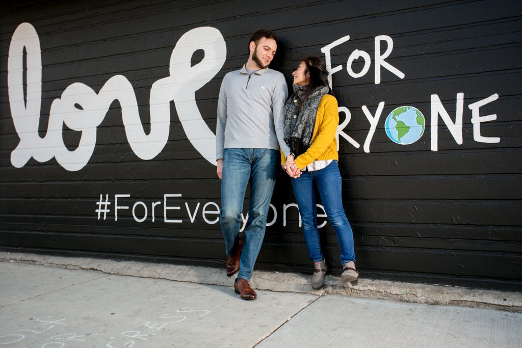 A young couple holds hands and smiles adoringly at one another against a graphic painted wall on a city street.