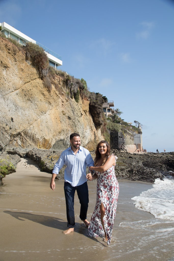 A newly engaged couple walk and laugh playfully on the beach as the water starts to roll in.