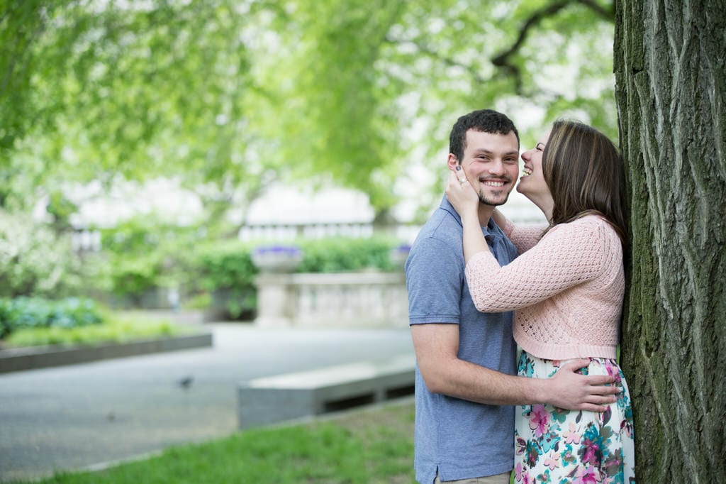 A newly engaged couple smiles and laughs with one another against a large tree in the park.