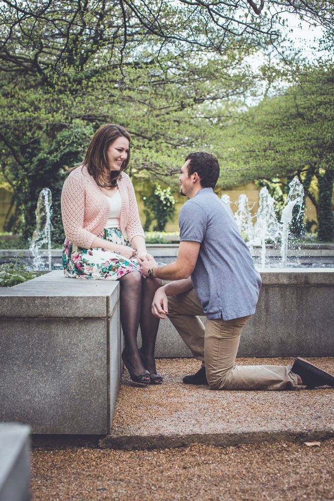 A man kneels on one knee to propose to a woman whilst surrounded by an intricate fountain in a park.