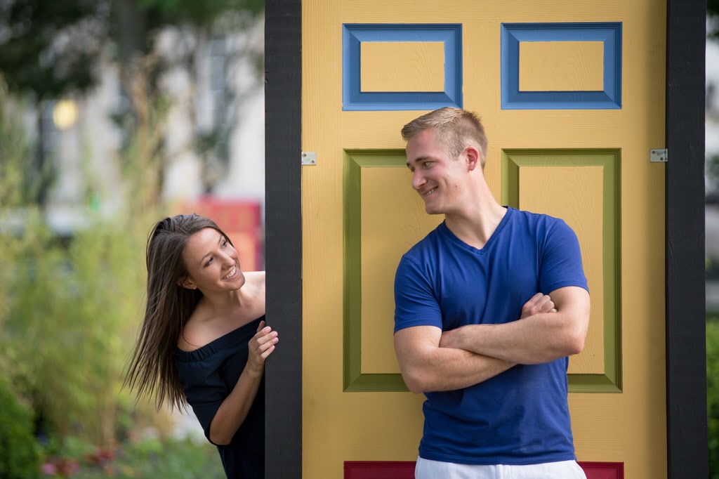 A newly engaged woman playfully peeks from behind a standalone doorway at her soon-to-be husband.