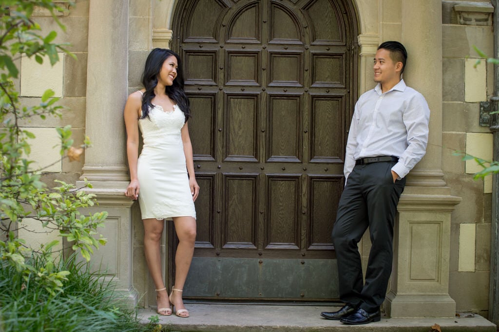 A newly engaged couple stands on either side of an old Victorian style door surrounded by sandstone bricks.