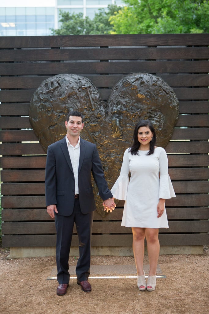 A newly engaged couple holds hands and smiles in front of a large heart sculpture.