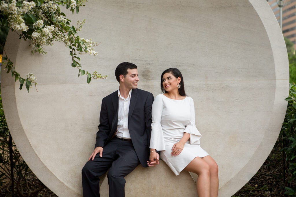 A newly engaged couple sit and hold hands in front of a large, circular sculpture.