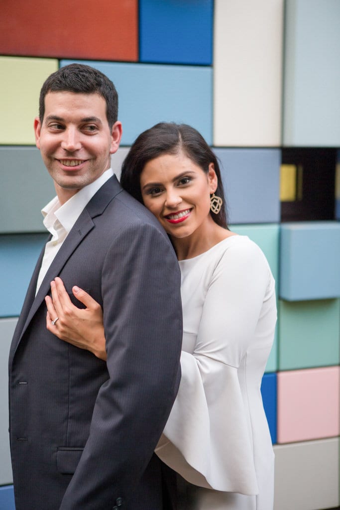 A newly engaged woman embraces her fiance from behind and lays her head on his back in a portrait style photo. The photographer captures the full, red smile of the woman as the man looks behind them with a grin of his own. Professional photography, wedding photography and engagement photography posing ideas.