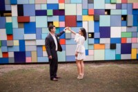 eivans_gallery_engagement_photos_189 A newly engaged couple smiles and dances in front of a multi-colored, geometric design wall. The woman wears a bell-sleeved, ivory skater dress with taupe, suede, peep-toe wedges.