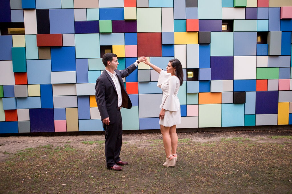 A newly engaged couple smiles and dances in front of a multi-colored, geometric design wall. The woman wears a bell-sleeved, ivory skater dress with taupe, suede, peep-toe wedges.