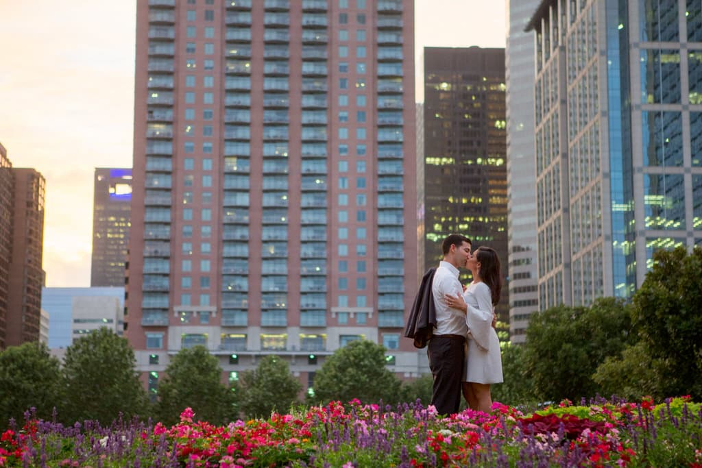 A newly engaged couple shares a kiss in the middle of a colorful city garden surrounded by skyscrapers.