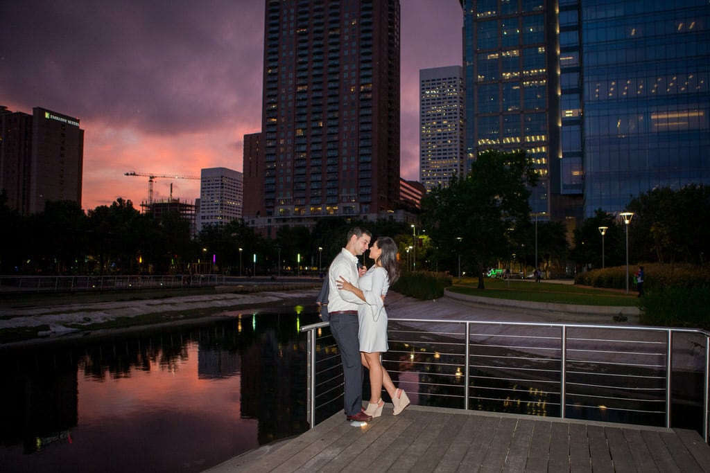 A couple holds one another firmly as the sun sets between the skyscrapers of the city. A small body of water next to them reflects the twilight skies and surrounding buildings.