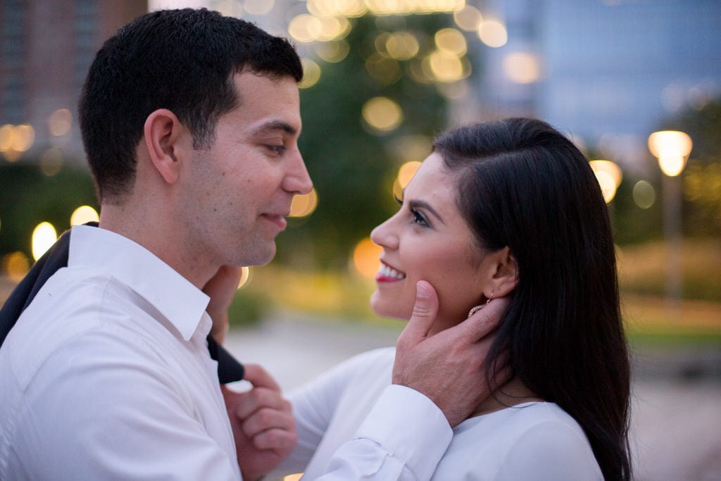 A couple stands in a city park. They gaze at one another as the woman smiles up at her soon-to-be husband and he cradles her head gently.