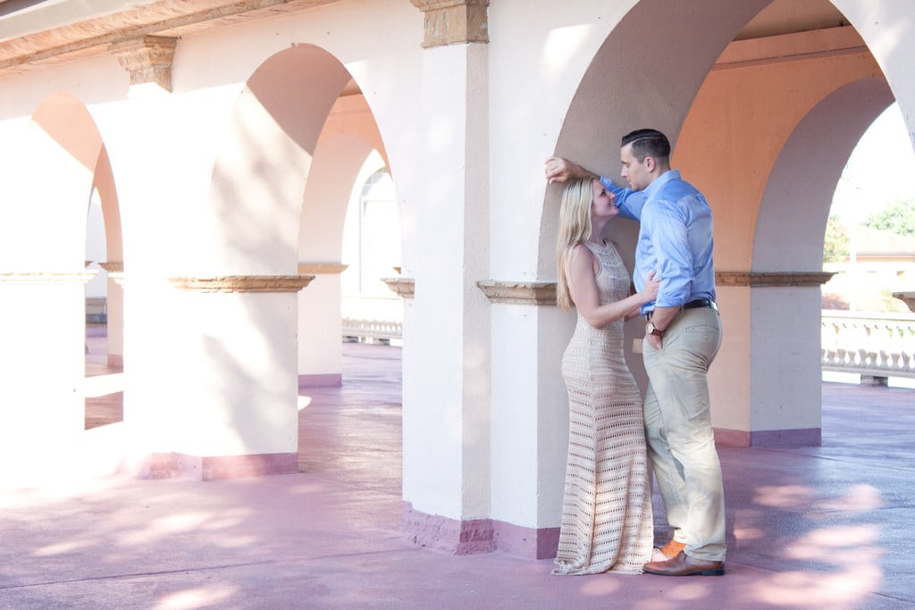 A couple stands, embracing one another beneath a sand-colored outdoor archway on a sunny day. The woman wears a sand-colored eyelet maxi dress matching the surroundings perfectly. The man stands protectively but adoringly over his fiance.