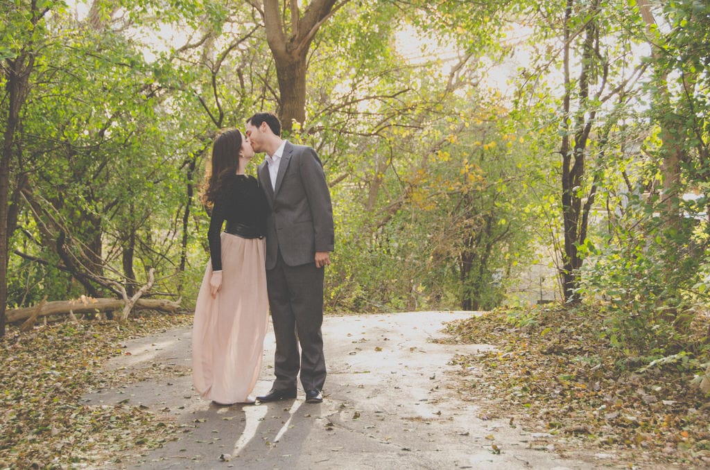 A young couple shares a kiss on a forest trail for their engagement shoot. The woman wears a flowy, pink chiffon maxi skirt with a black, leather waist belt.