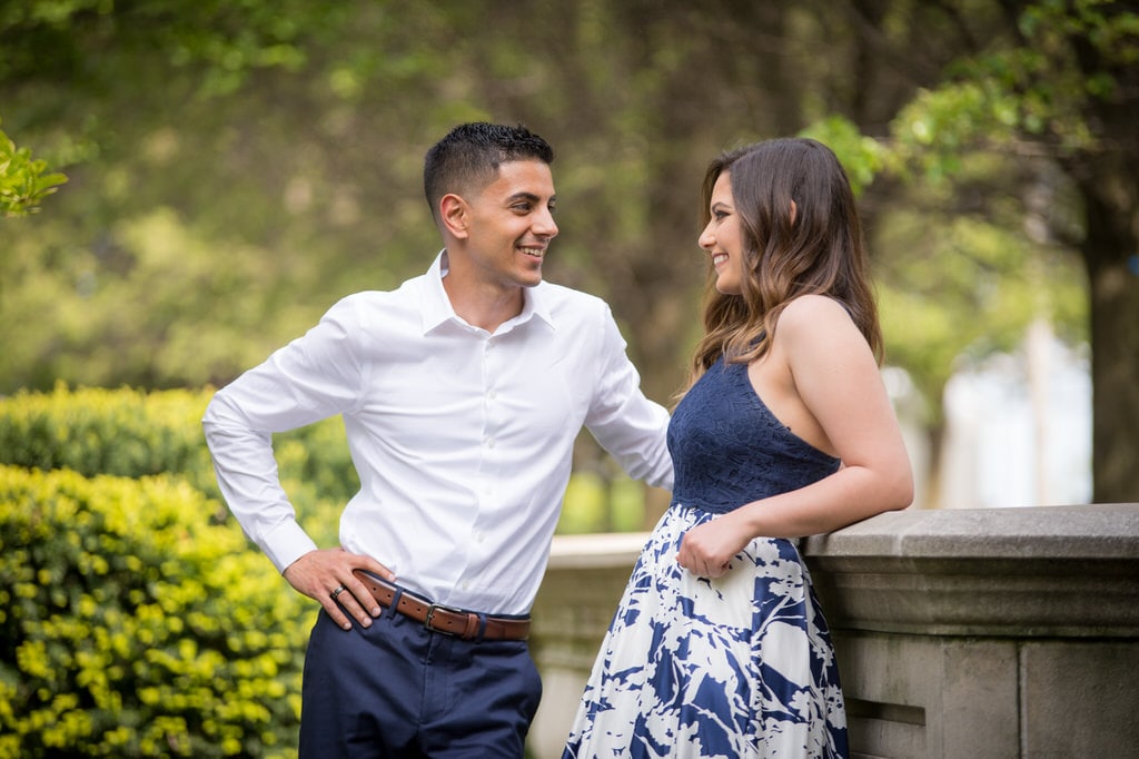 A man is leaning against a stone wall in a park, looking at his fiance as they smile at each other.