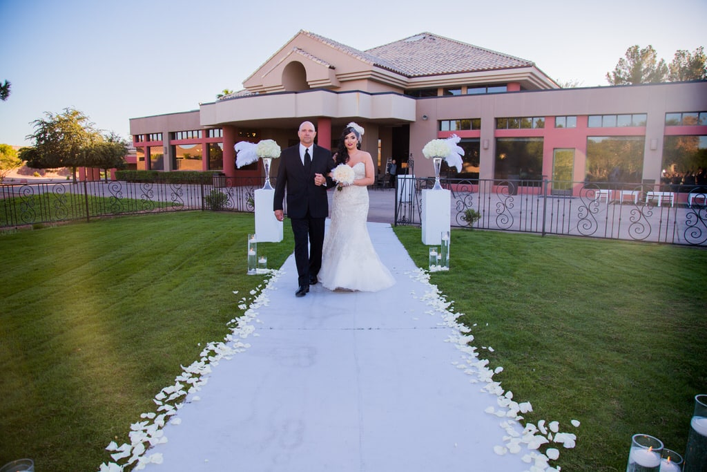 A father walks his daughter down the aisle lined with white rose petals. There are floating tea light candles placed in the entryway. She is carrying a white rose and feather bouquet and matching birdcage visor with netting.