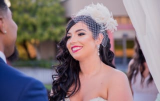 New Bride smiles adoringly at her new husband during the ceremony. The Bride wears an ivory, feathered birdcage visor with netting.