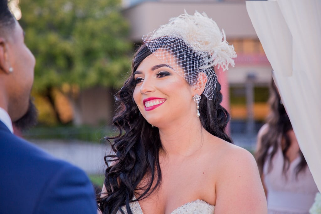 New Bride smiles adoringly at her new husband during the ceremony. The Bride wears an ivory, feathered birdcage visor with netting.